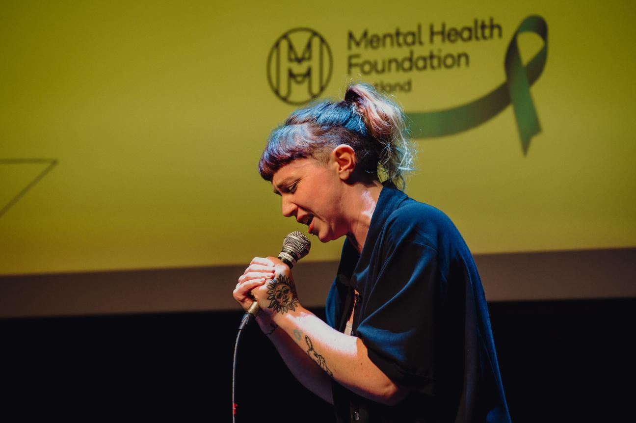 A person performing with a microphone, in front of the MHF Scotland logo