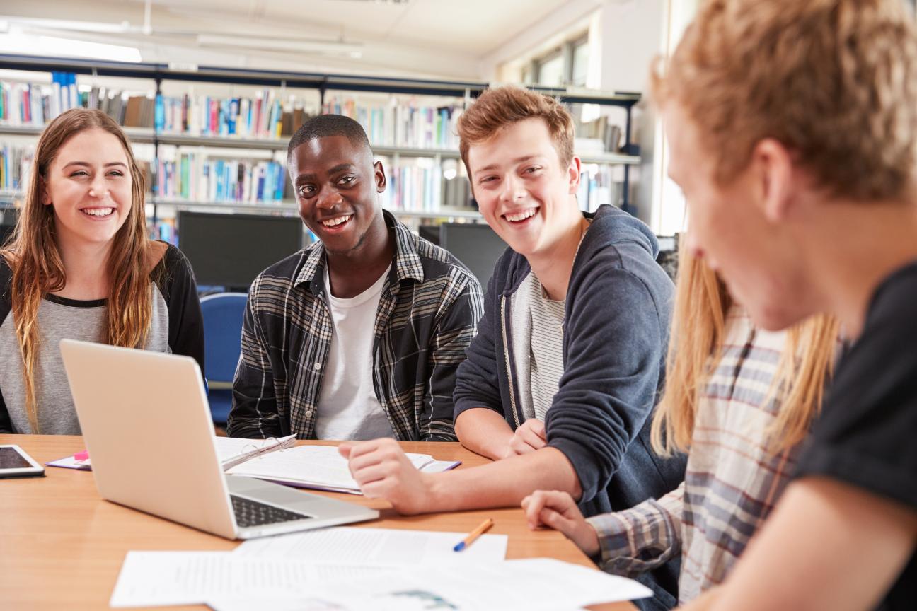 A group of smiling students sitting around a table in a college library talking