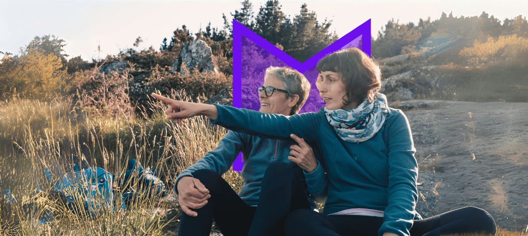 Two women sit in nature, looking to the left of frame where one of them is pointing.