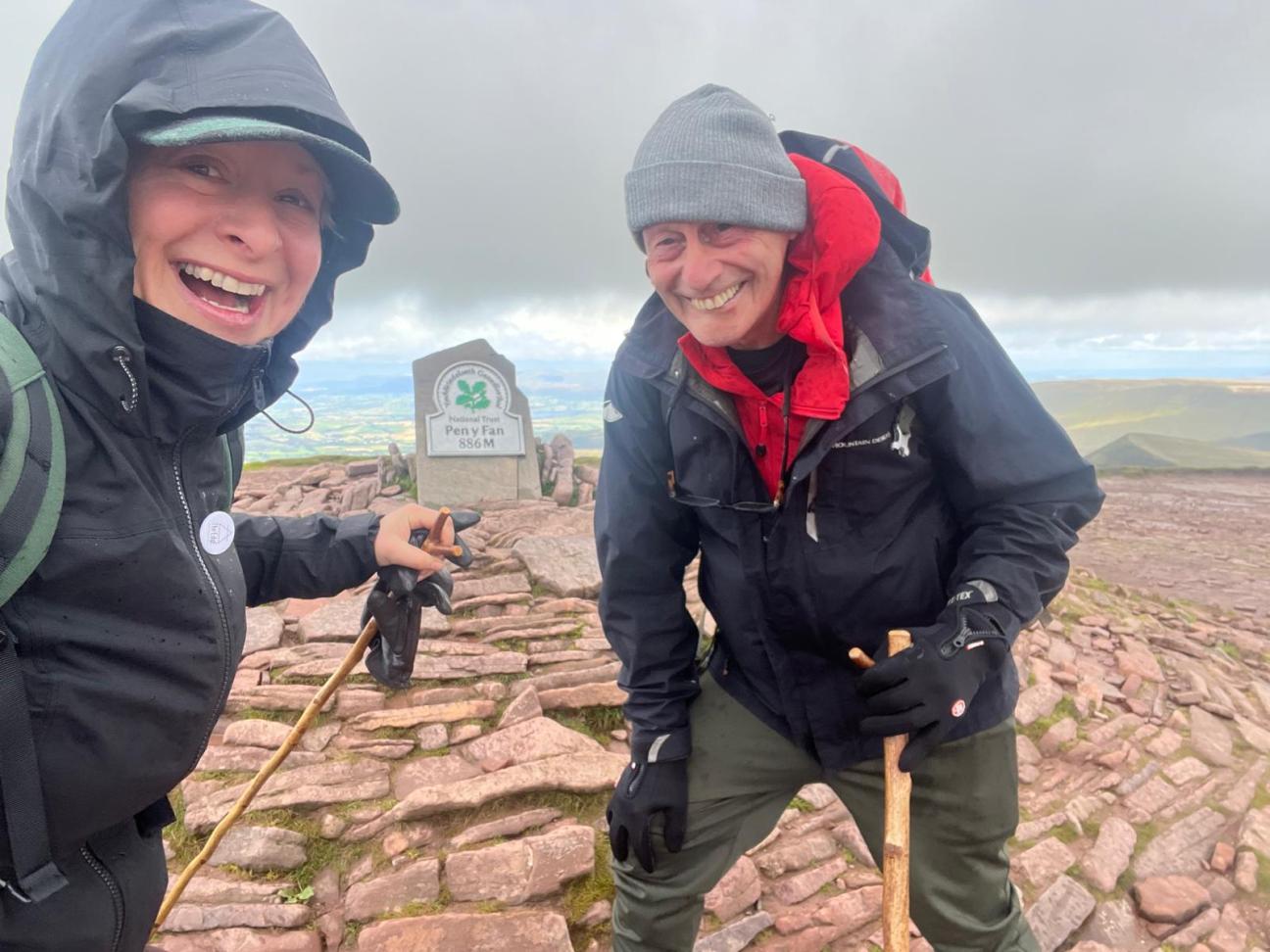 A couple stood smiling in the wind at the top of Pen Y Fan mountain