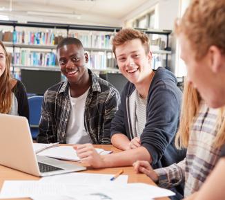 A group of smiling students sitting around a table in a college library talking