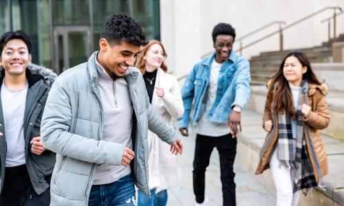 A group of five smiling young adults walk outdoors, bundled in winter jackets. The mood is joyful, with steps, glass building, and urban setting in background.