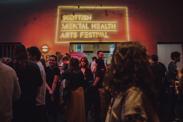 Photo from Scottish Mental Health Arts Festival, featuring the crowd and the logo in a neon sign
