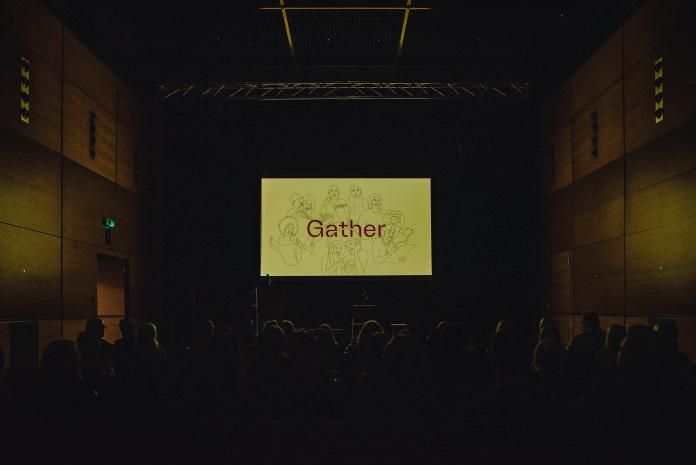 A crowd looking towards a screen showing the SMHAF 2022 graphic for the theme 'gather'