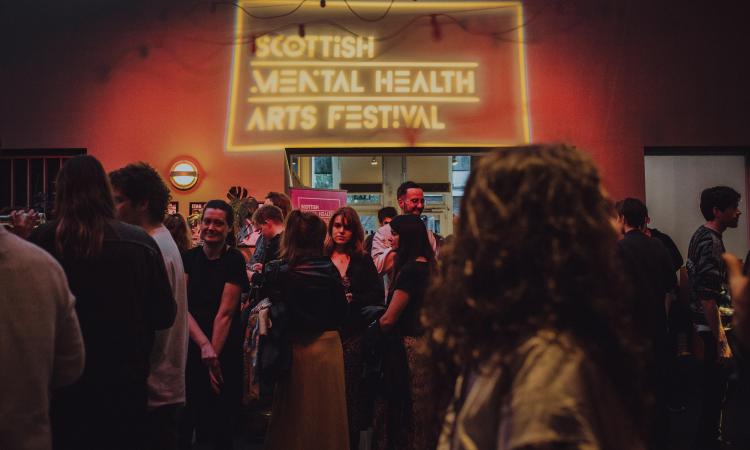 Photo from Scottish Mental Health Arts Festival, featuring the crowd and the logo in a neon sign