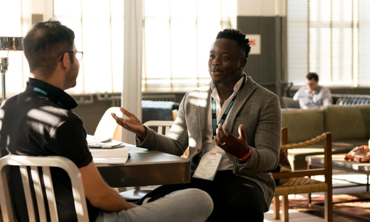 Two men sitting talking at a table. One has his back to us, the other is gesturing with his arms.