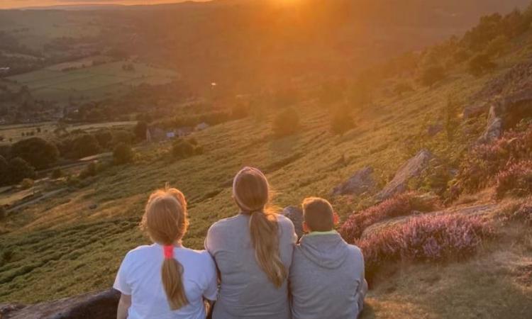 A woman is sat between two children with their backs to the camera, looking out at a sunset over green hills.