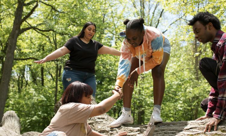 Four people outdoors, two helping the other climb over a log. One wears a tie-dye hoodie and the other a black shirt. Supportive and collaborative scene.