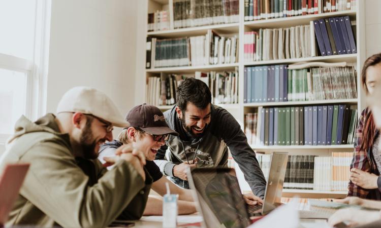A group of four people are laughing and collaborating at a table with laptops in a bright library, conveying a lively and cheerful atmosphere.