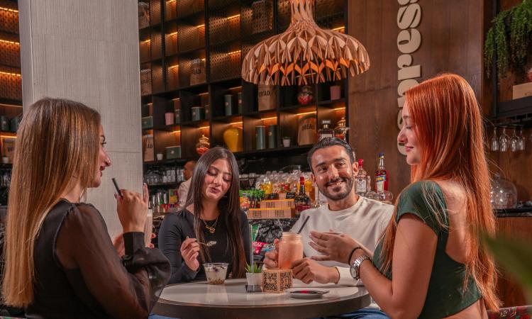A group of friends shares drinks and laughter around a cafe table. Warm lighting, colorful chairs, and a modern interior create a cozy, cheerful atmosphere.