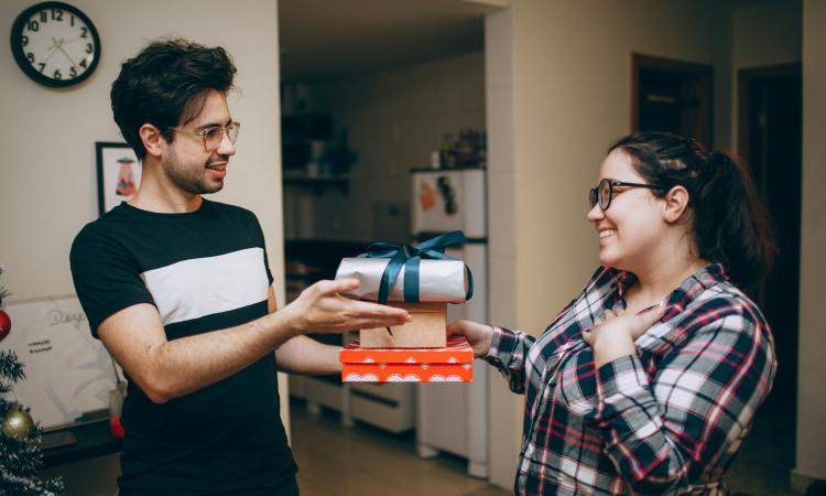 A man in a black shirt gives a gift to a smiling woman in a plaid shirt. The setting is a cozy kitchen with a festive feel.