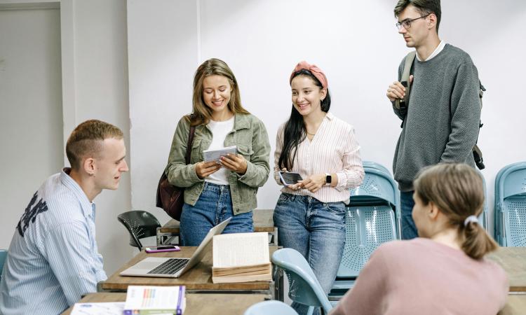 A group of five students in casual attire gather around a table in a bright classroom, engaging in conversation and smiling, creating a positive, collaborative mood.