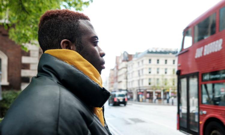 A Black man waiting for a bus on the side of a road, you can see a London bus on the road passing by.
