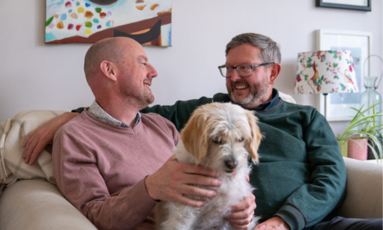 A couple sit close together on a sofa with their pet dog talking and smiling