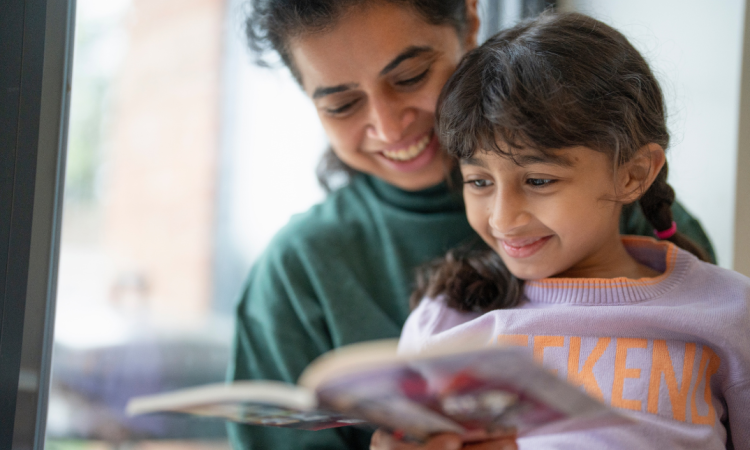 A woman reads a book to her young daughter, both are smiling