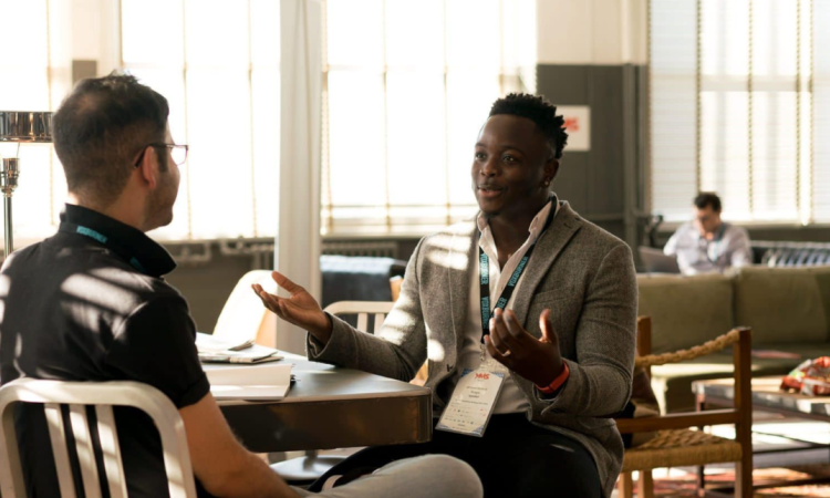 A Black man wearing a suit and lanyard sits at a table, talking to another man. 