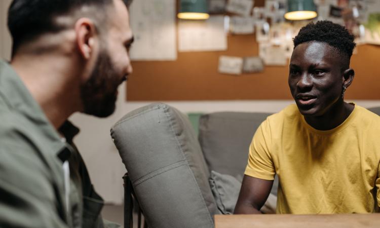 A young Black man in a yellow top sits on a sofa talking to another man in the foreground. 