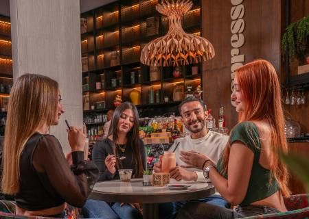 A group of friends shares drinks and laughter around a cafe table. Warm lighting, colorful chairs, and a modern interior create a cozy, cheerful atmosphere.