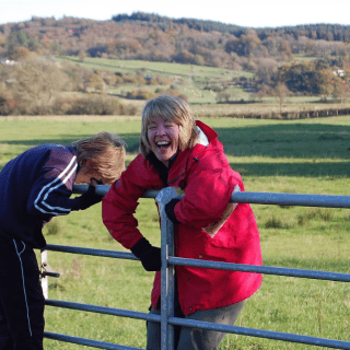 A mum and son hold onto a gate beside a field in the countryside, both are laughing