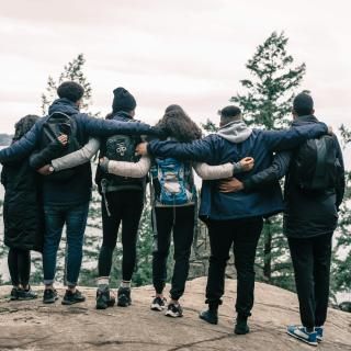 Six friends stand arm-in-arm on a rocky cliff edge, overlooking a forest and distant water. The scene conveys warmth, unity, and adventure.