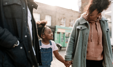 A Black mother and her child walk hand in hand on a street, smiling.