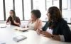 Three women sitting at a table in a meeting, in front of a couple of them on the desk are notebooks